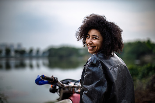 Beautiful girl with afro hair, wearing biker jacket, riding a dirt bike reaching the lakeside in Tay Ho, Hanoi, Vietnam