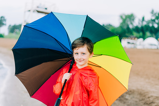 Portrait of a smiling school boy with rainbow umbrella in the park. Kid holds colourful umbrella on his shoulder. Cheerful child in a red raincoat holding multicolor umbrella outdoors.