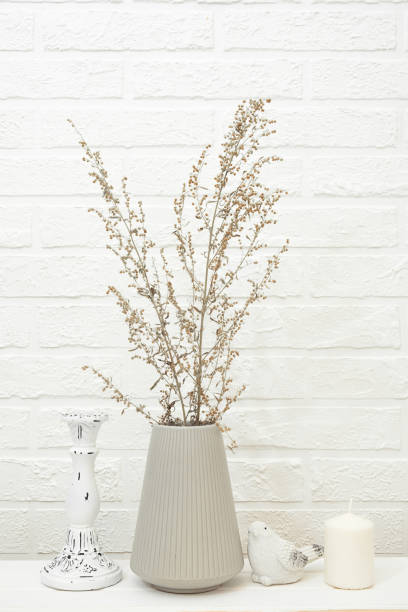 istock Bouquet of dry grass branches in modern gray ceramic vase and decorative items on white table against white brick wall. Minimalistic composition in neutral tones in Scandinavian style. 1425846948