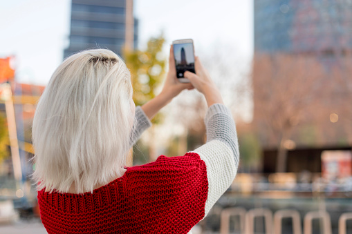 Young blonde girl in red and white sweater taking picture with smartphone on the city