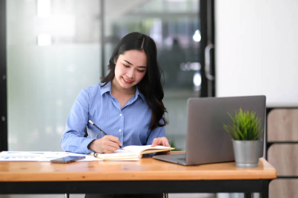 istock Charming asian businesswoman working on laptop and taking note while sitting at the table in office. 1425853108