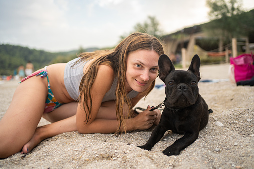 Portrait of woman with French Bulldog at beach, she's looking at camera and smiling
