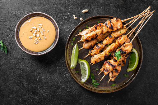 Chicken grilled satay skewers served with lime and peanut sauce on black, top view. Asian thai style food.