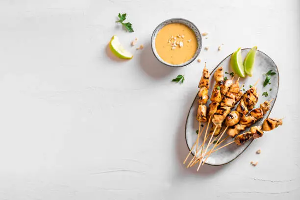Chicken grilled satay skewers served with lime and peanut sauce on white, top view, copy space. Asian thai style food.