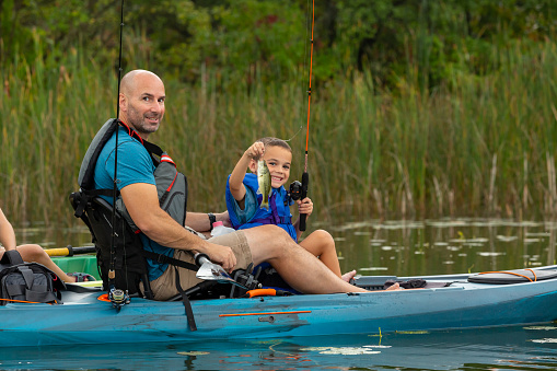 Close-up of a young boy in a kayak with his dad. The boy is holding up a smallmouth bass he just caught while kayak fishing.