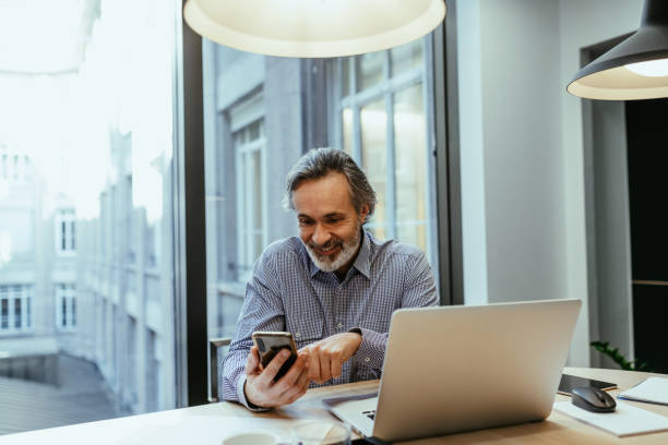 istock Gray hair employee working in office, looking at smart phone while using mobile app 1425872243
