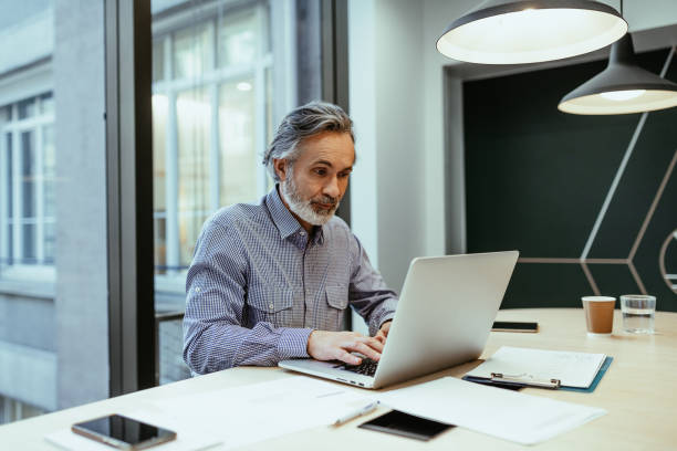istock Silver hair employee working in office, looking at computer and smiling 1425872358