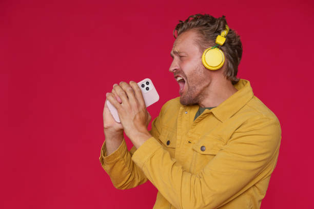 istock Handsome man 30s singing enjoying his favorite song or track using phone and wireless headphones wearing denim yellow jacket isolated on red background. Joyful man sing while listen music 1425880421