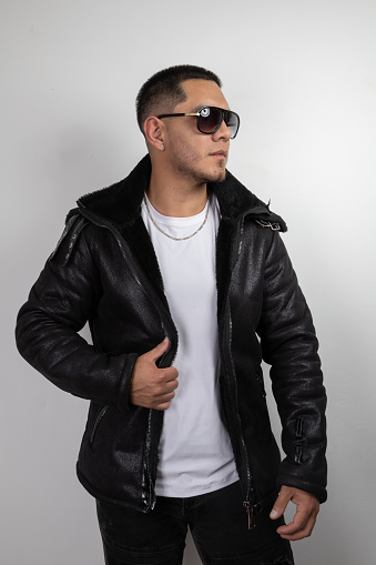 attractive latin young man modeling trendy modern clothes, he wears a leather jacket, white t-shirt, sunglasses and necklace, beauty in studio