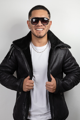 young model with short hair wearing sunglasses, leather sweater, white t-shirt and necklace while smiling, urban fashion and style, latin man in studio