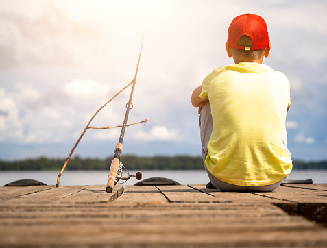 Young Hispanic and African American fishing buddies walk to the lake or pond to fish together. An Hispanic boy is carrying a fishing pole and tackle box. An African American boy is holding a fishing pole and net in the background.