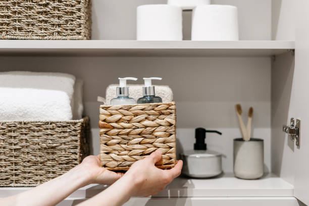 Woman put wicker box with cosmetics products in bathroom closet Organization of space in the bathroom cabinet. Cropped view of woman putting wicker box with bath sponge, shampoo, soap dispenser bottle and other cosmetics products in closet paper dispenser stock pictures, royalty-free photos & images