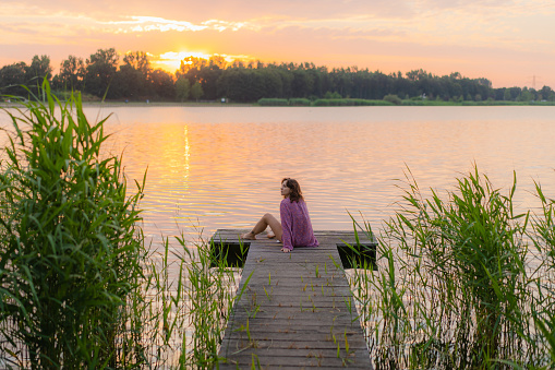 Young Caucasian woman sitting on pier at the lake in summer at sunset