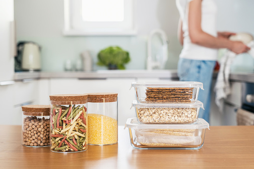 selective focus on food containers with pasta, grains, chickpeas and oats on wooden table with blurred kitchen background, storage in glassware kitchenware