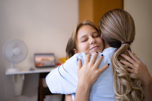 Teenage daughter having her eyes closed, hugging her mother and bonding with her at home