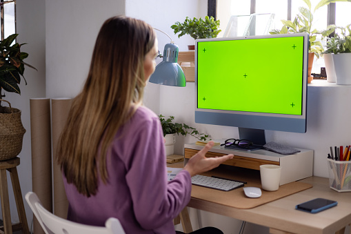 Young adult female freelancer sitting at her desk in her home office, casually working from home using a desktop PC with a chroma key and having a web conference with her colleagues