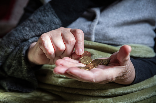 senior adult or pensioner is sitting with winter clothes indoors under a warm blanket, counting some coins, concept energy crisis