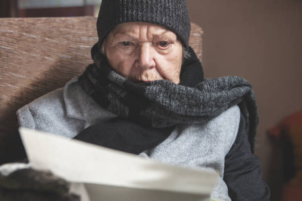 senior adult or pensioner is sitting with winter clothes indoors under a warm blanket, reading a letter or bill, concept energy crisis