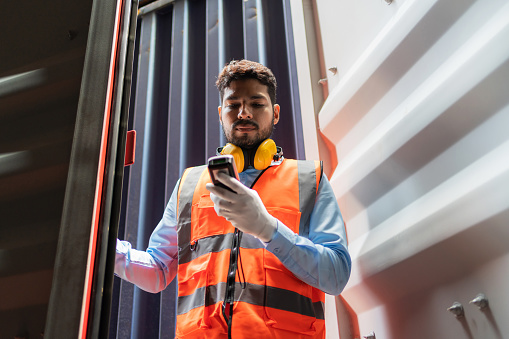 Dock worker man in safety vest holding barcode scanner inspector the quality of container at shipping yard. Logistic and Transportation, Global Business concept. Cargo freight ship for import export