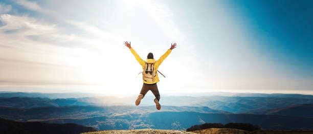 Happy hiker jumping on the top of the mountain at sunset - Man with backpack and arms up standing on the cliff - Happiness and travel concept stock photo