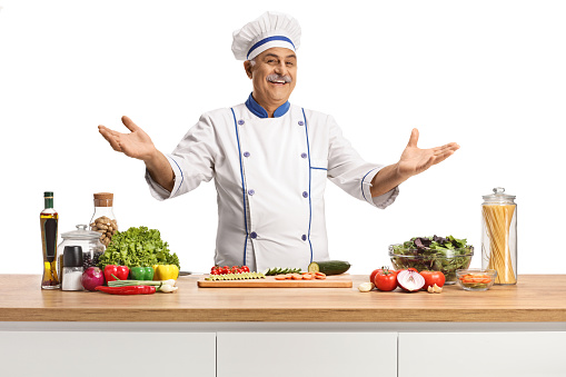 Cheerful mature male chef posing behing a kitchen counter with vegetables isolated on white background