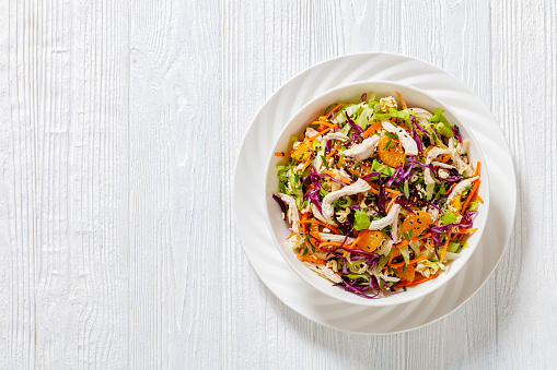 Chinese Chicken Salad with shredded chicken meat, mandarin oranges, crunchy noodles, red cabbage, napa cabbage, carrots, green onions in bowl, flat lay, free space