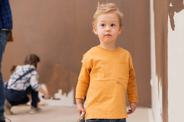 um menino loiro, vestido com uma blusa laranja, está em uma sala onde os reparos estão acontecendo e está pensando em algo. ao fundo, sua irmã mais velha está pintando a parede marrom - brown hair caucasian little boys men - fotografias e filmes do acervo