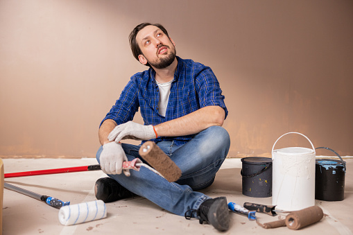37-year-old man with a beard and mustache wearing blue jeans checkered shirt white T-shirt and black sneakers. Is sitting on ground in middle of cans of warb. He holds paint roller in hand.