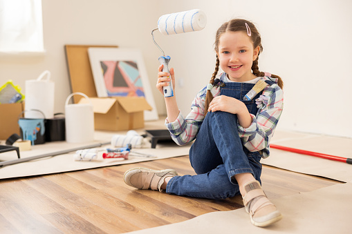 Girl sitting on floor wearing blue denim overalls checkered shirt and beige sneakers. Kid holds in right hand paint roller for painting walls. In background things to be repaired buckets of paint.