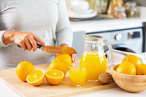 Close up view of woman's hands cutting oranges in halves with a knife. Whole and halved oranges, jug and drinking glass filled with squeezed orange juice are on a cutting board. High resolution 42Mp indoors digital capture taken with Sony A7rII and Sony FE 90mm f2.8 macro G OSS lens