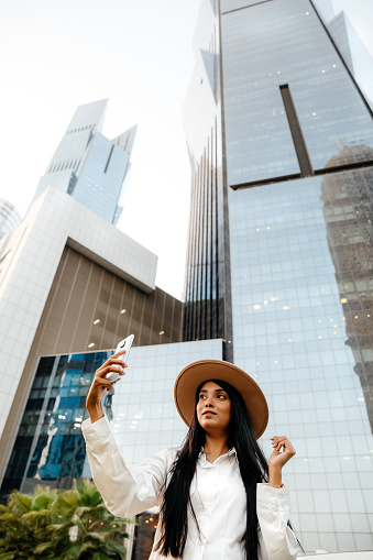 Smiling woman solo traveller with trendy look making selfies on mobile phone, skyscrapers on a background in Doha, Qatar