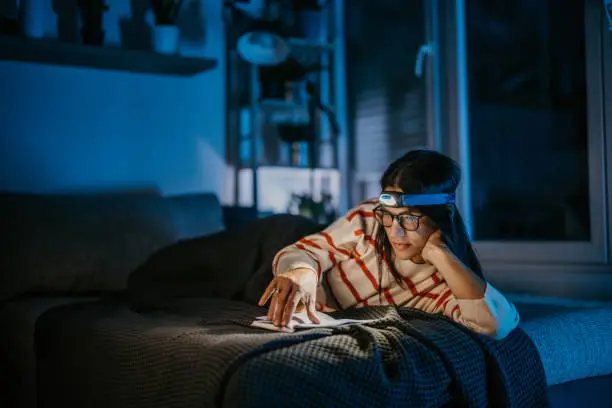 Woman covered in blanket lying in bed at home at night, she is reading a book while wearing headlamp, because there is no electricity in the apartment.