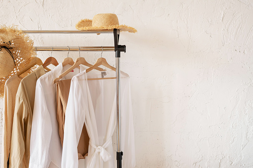 Trendy capsule wardrobe in beige and white on a rail rack with straw hats. Copy space