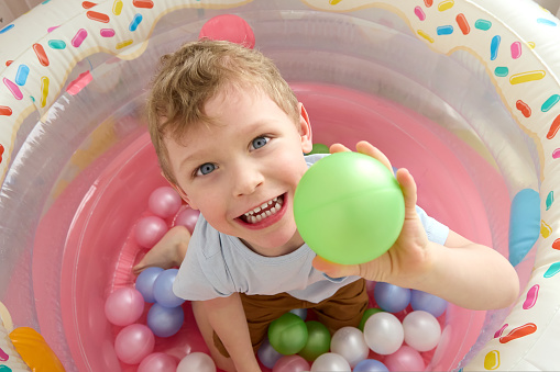 Blond haired little boy throws colorful plastic balls while standing in a dry children's pool in the children's room. A bright photo of a happy child smiling while playing, looking at the camera.