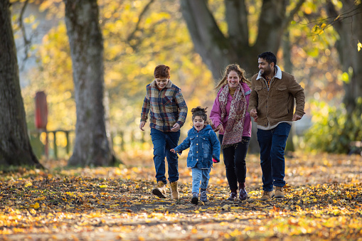 A family spending the day together outdoors in Hexham, North East England during Autumn. They are running towards the camera on a footpath covered in fallen leaves while laughing and smiling with each other.