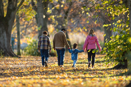 A family spending the day together outdoors in Hexham, North East England during Autumn. They are walking away from the camera on a footpath covered in fallen leaves and the mother and father are holding hands with their daughter.