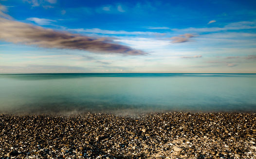 Dreamy, calm image depicting a long exposure with silk smooth sea water, a pebble beach in the foreground, and motion blur of a cloudscape and blue sky beyond.