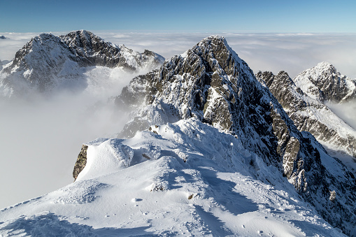 Looking across to the top of a snowy, rocky mountain range, with a layer of cloud cover not too far below the peaks. There is mist just above the clouds and blue sky and sunshine on the landscape.