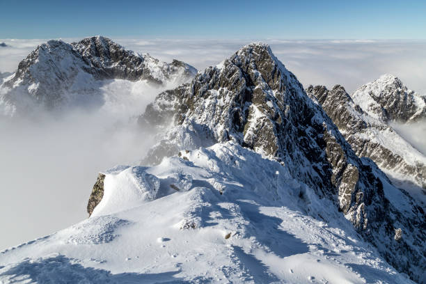 View of dramatic, rocky, snowy mountain range peaks with mist and clouds, Lomnicky Stit, High Tatras, Slovakia, European alps. Looking across to the top of a snowy, rocky mountain range, with a layer of cloud cover not too far below the peaks. There is mist just above the clouds and blue sky and sunshine on the landscape. high tatras stock pictures, royalty-free photos & images