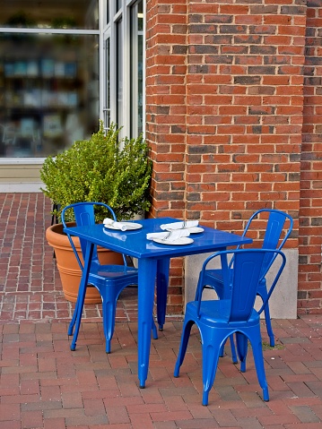 Outside dining setting on brick street in downtown Lexington Massachusetts. Table place setting reside on contrasting blue table and chairs with red brick background. Copy space and store window in background.