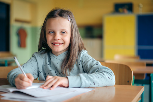 schoolgirl sitting at classroom desk and writing in her workbook. Elementary schoolgirl studying in classroom.
