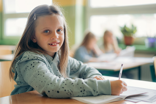 schoolgirl sitting at classroom desk and writing in her workbook. Elementary schoolgirl studying in classroom.
