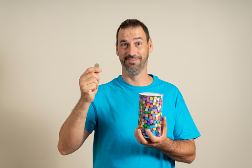 Smiling latin man with a beard dressed in a blue t-shirt putting a euro coin in a metal piggy bank, trying to save so he can travel on vacation. Isolated on beige studio background.