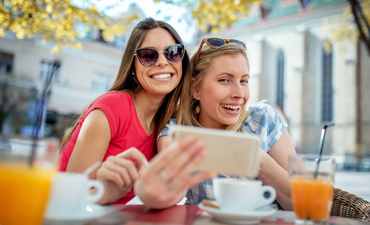 Young girls sitting in the cafe and talking with friends via video call on smartphone