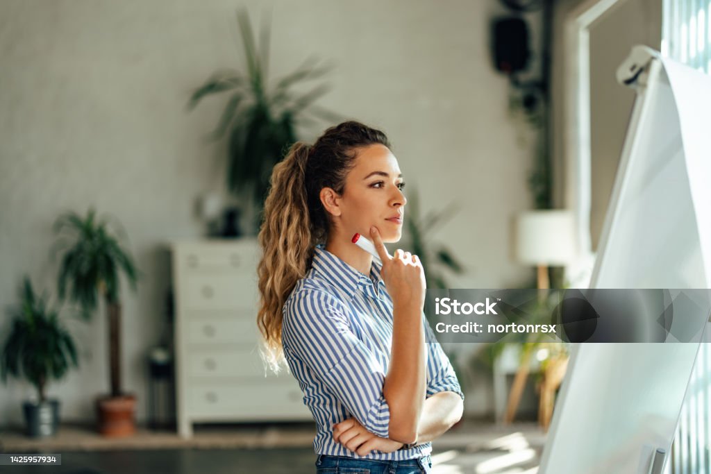Concentrated girl looking at the white board, holding a marker. Blonde female worker looking thoughtful, creating plans, writing on the board. Contemplation Stock Photo