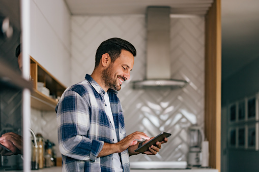 Smiling adult man holding a tablet, using it while being in the kitchen.