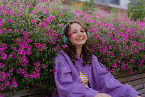 Young Caucasian woman in purple coat  listening to music in headphones on the background of purple flowers