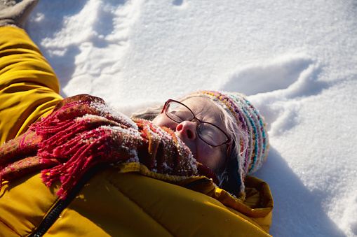 senior woman, 67 years old, lying in snow, wearing orange jacket and colorful cap, enjoying a sunny summer day