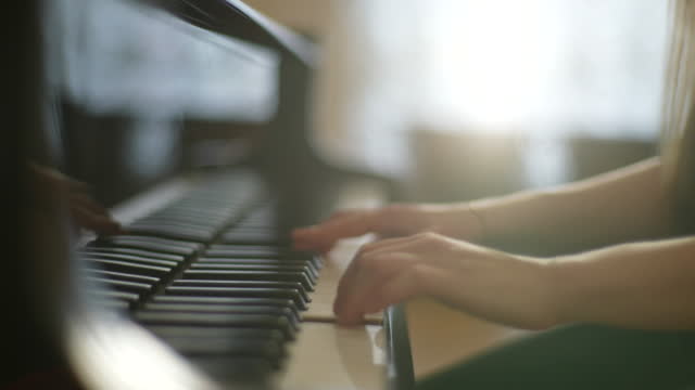 Close-up tracking shot of unrecognizable talented young woman pianist playing gentle music on beautiful grand piano sitting on background of window.