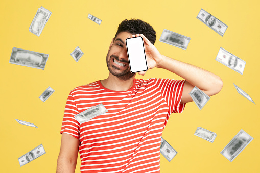 Online investment, winner overjoyed rich man and money earning. Satisfied guy covering his eye with mobile empty display and rejoicing in his wealth. Indoor studio shot isolated on yellow background.